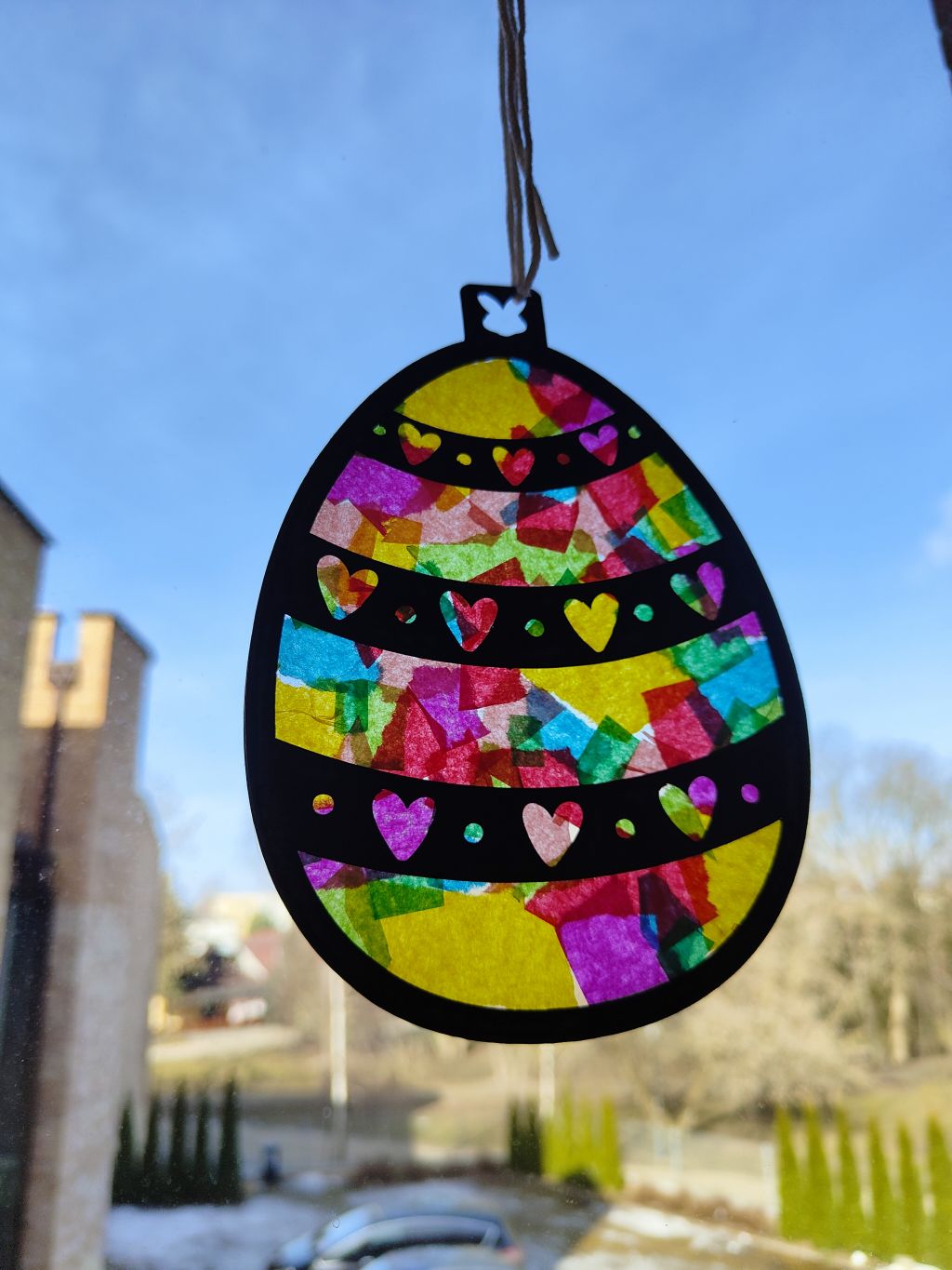 Zbliżenie na gotową ozdobę wielkanocną w kształcie jajka, zawieszoną na cienkim sznurku na tle okna. Ozdoba ma czarną ramkę z wyciętymi wzorami serc i kropek, a jej wnętrze wypełnione jest kolorową, półtransparentną bibułą w odcieniach żółci, różu, czerwieni, zieleni i błękitu. Przez witraż prześwituje światło dzienne, a w tle widać rozmazany krajobraz z niebem, drzewami i fragmentem budynku.