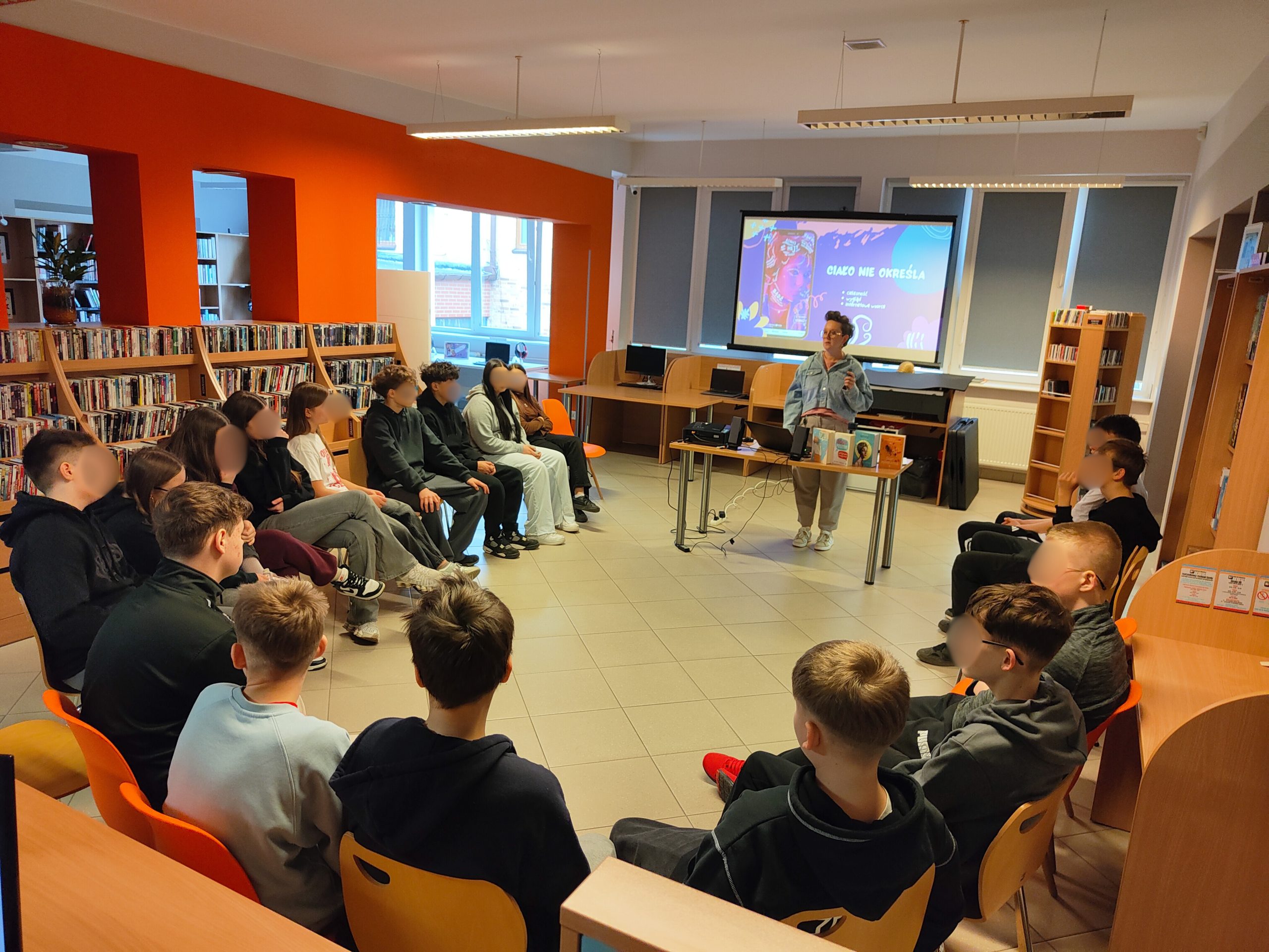 Grupa osób siedząca w półkolu przed ekranem z prezentacją w nowoczesnej bibliotece.