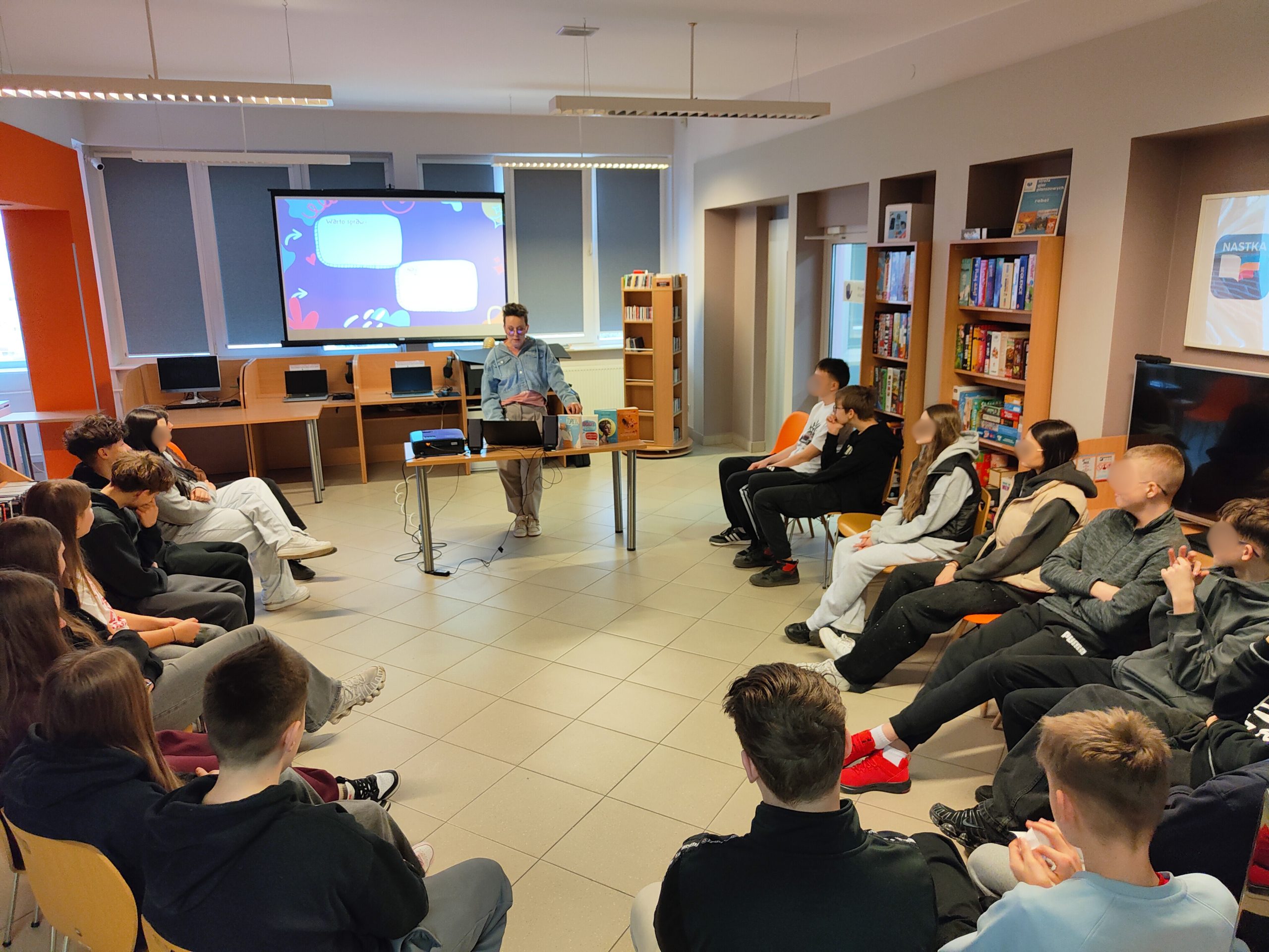 Prowadząca warsztaty stoi przy stole z laptopem i książkami, przemawiając do grupy młodzieży.