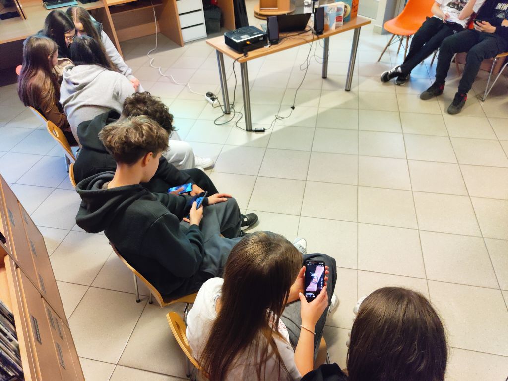 Młodzież siedząca w rzędzie w bibliotece, każda osoba korzysta z własnego smartfona.