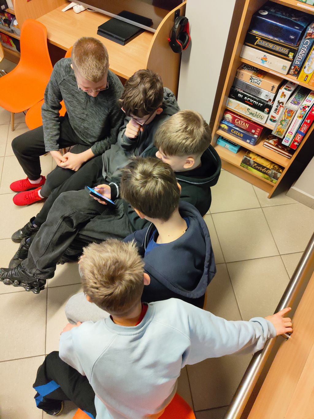 Grupa nastolatków siedząca blisko siebie i skupiona na ekranie jednego telefonu.