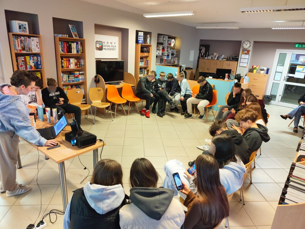 Widok na salę warsztatową z prowadzącą przy laptopie i młodzieżą siedzącą w kręgu.