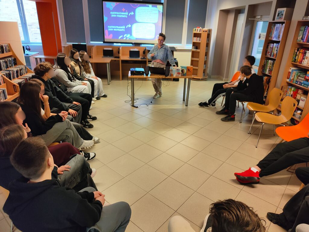 Uczestnicy spotkania siedzący w półkolu i słuchający wykładu w nowoczesnej bibliotece.