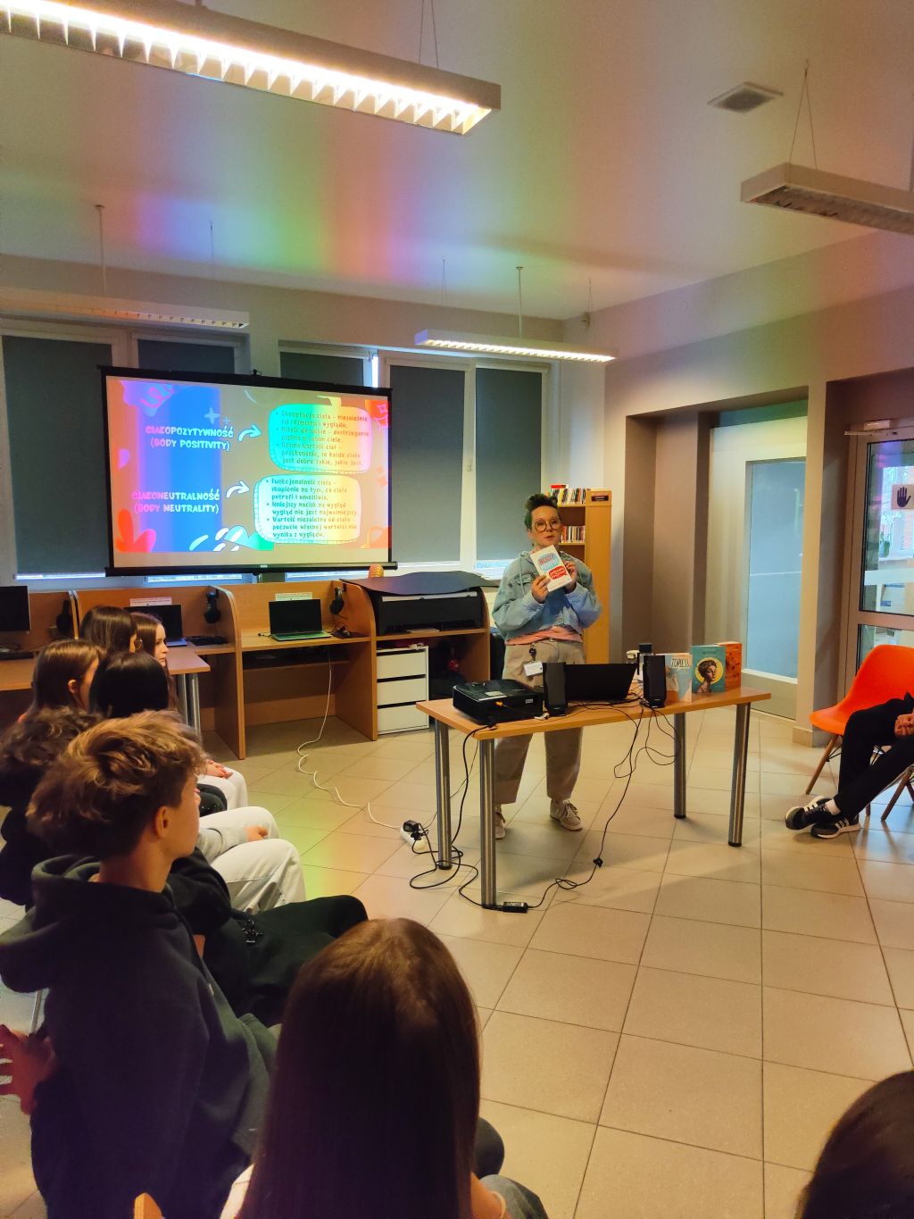 Prowadząca warsztaty trzymająca książkę „Body Positive” na tle ekranu z prezentacją.