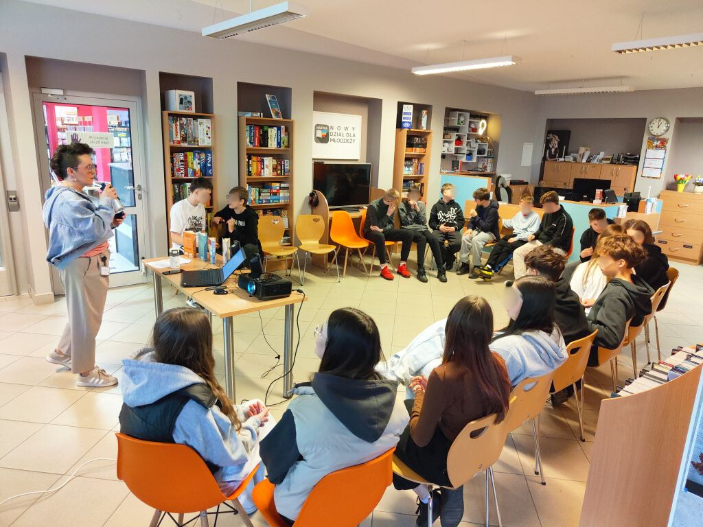 Grupa młodzieży siedząca w kręgu w bibliotece podczas spotkania z prowadzącą.