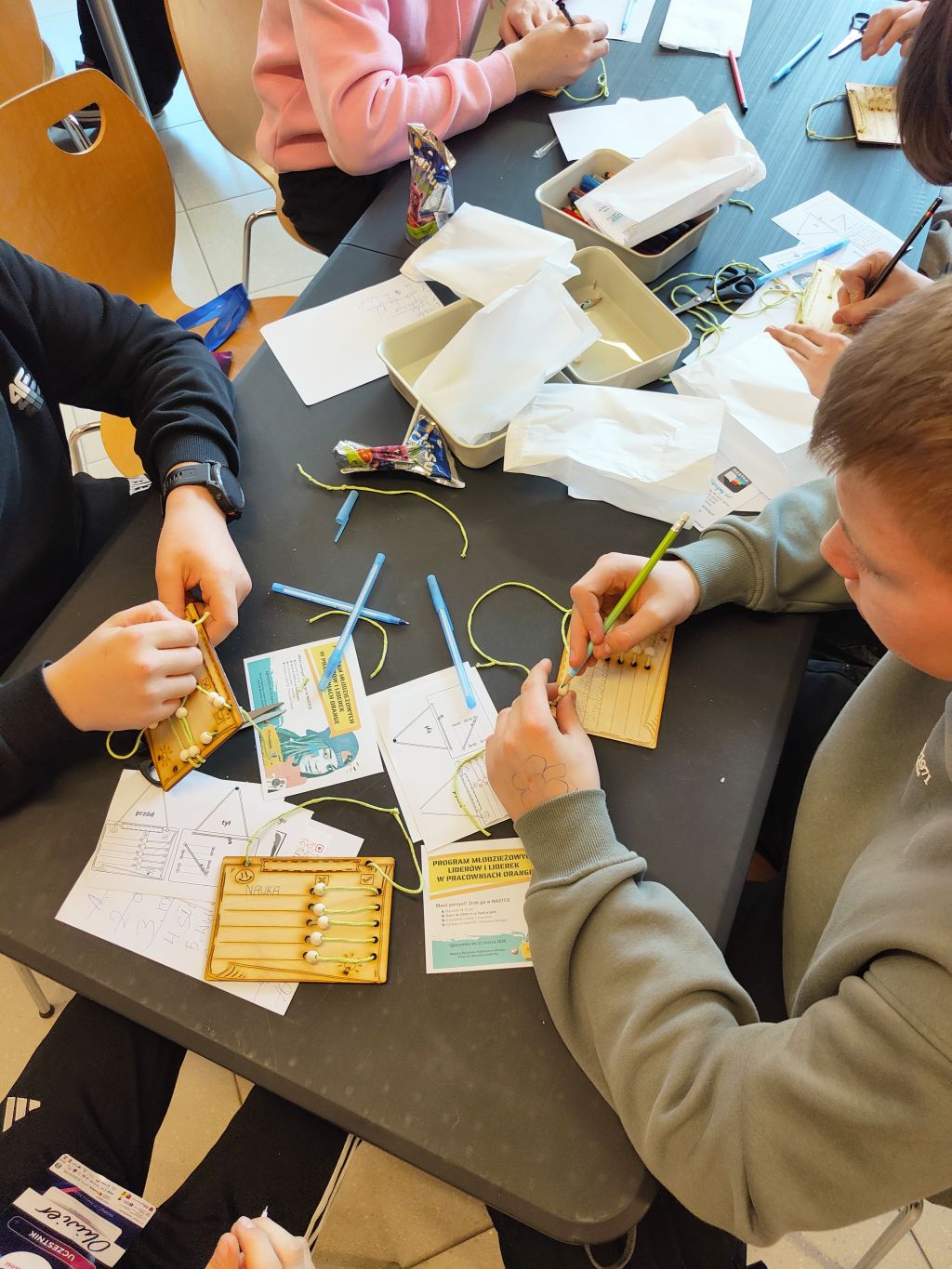 Uczestnicy warsztatów tworzą prace ręczne przy stole, korzystając z materiałów i instrukcji.