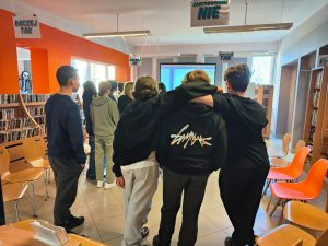 Trzech chłopców stoi tyłem do obiektywu, obejmując się ramionami i patrząc w stronę ekranu projekcyjnego. Jeden z nich ma na bluzie biały napis o ozdobnym kroju. W tle widać regały z książkami.