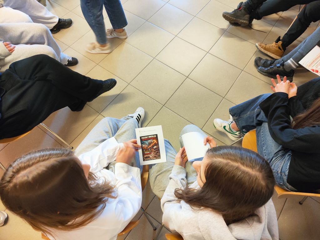 Widok z góry na dwie osoby siedzące obok siebie, które trzymają i wspólnie czytają ulotkę z grafiką przedstawiającą pszczoły i napisami w języku polskim.