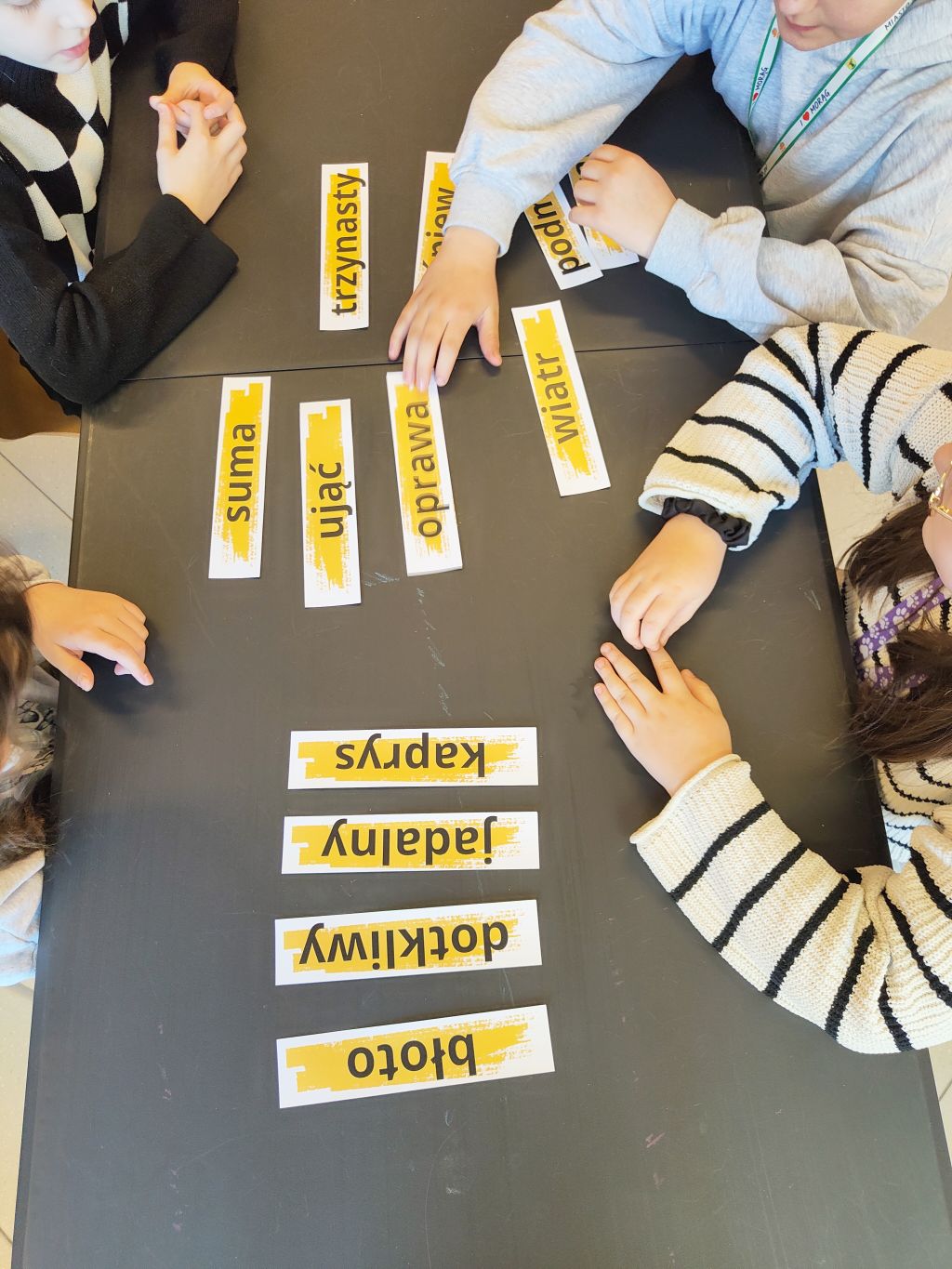 Dzieci siedzące przy stole porządkują żółte paski z polskimi słowami