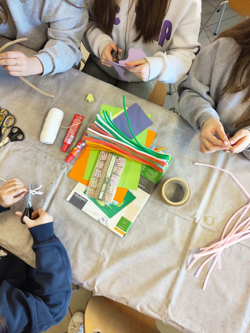 Widok z góry na stół warsztatowy, przy którym dzieci wycinają elementy z papieru. Na środku leży stos kolorowych drucików, papier kolorowy, kleje i taśma klejąca.