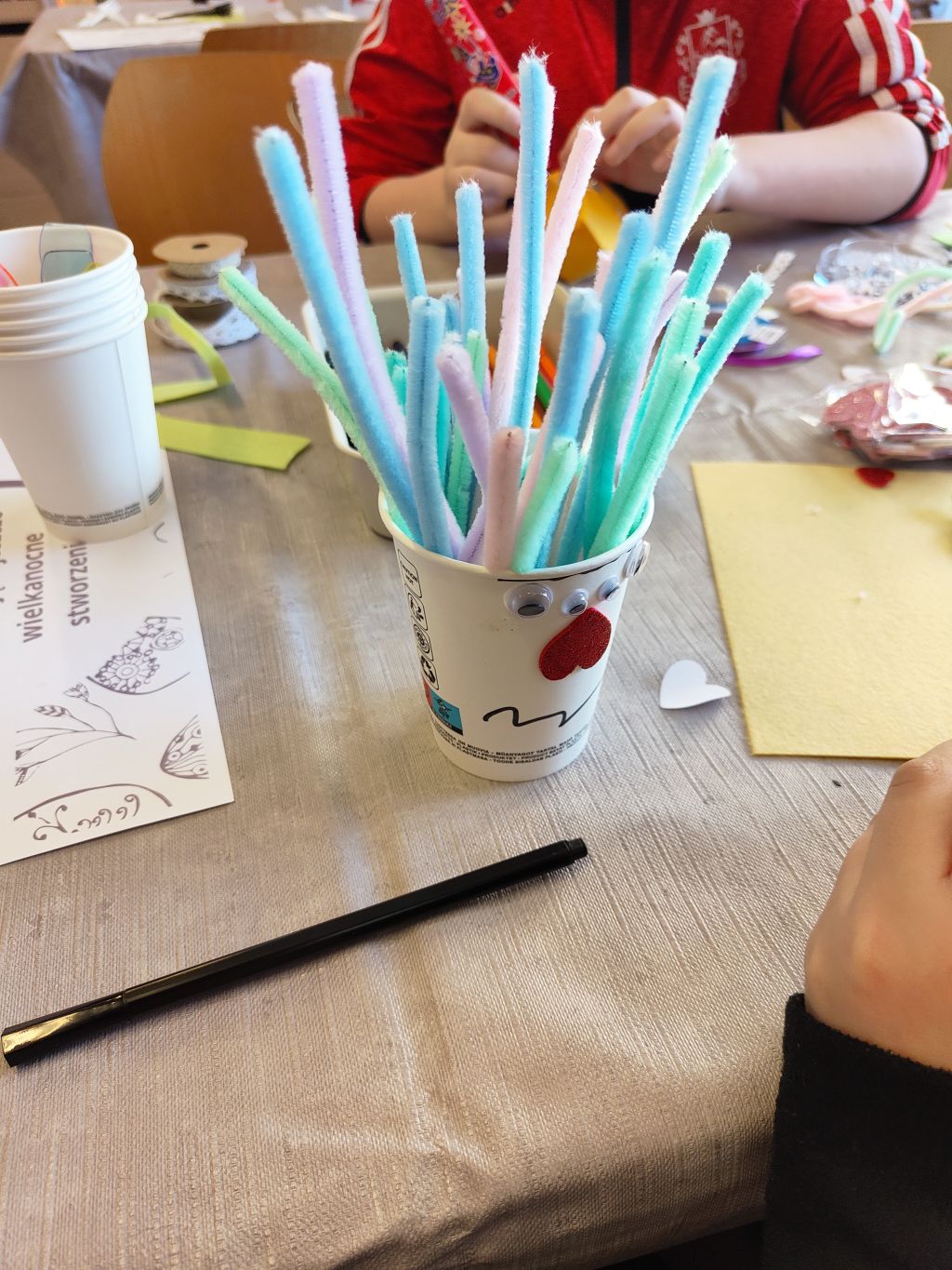 Biały papierowy kubek z przyklejonymi ruchomymi oczkami i serduszkiem, wypełniony pionowo kolorowymi drucikami kreatywnymi. W tle widoczne rozmazane postacie dzieci przy stole.