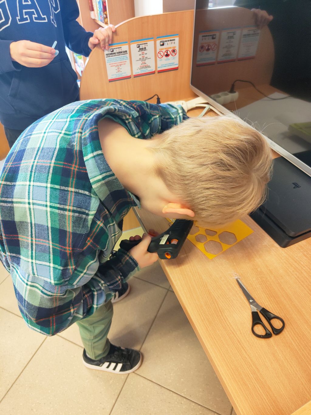Dziecko w koszuli w kratę używa pistoletu na klej na gorąco przy drewnianym blacie. Obok leżą nożyczki i żółty szablon z wyciętymi otworami.