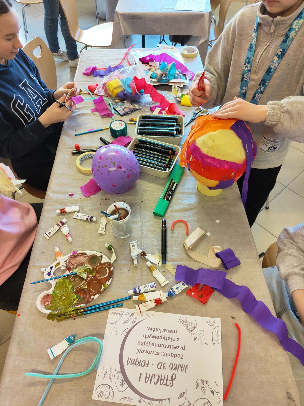 Dzieci przy stole tworzą prace plastyczne. Na pierwszym planie leży instrukcja, obok widać paletę z farbami, kolorową bibułę oraz oklejany papierem balon.