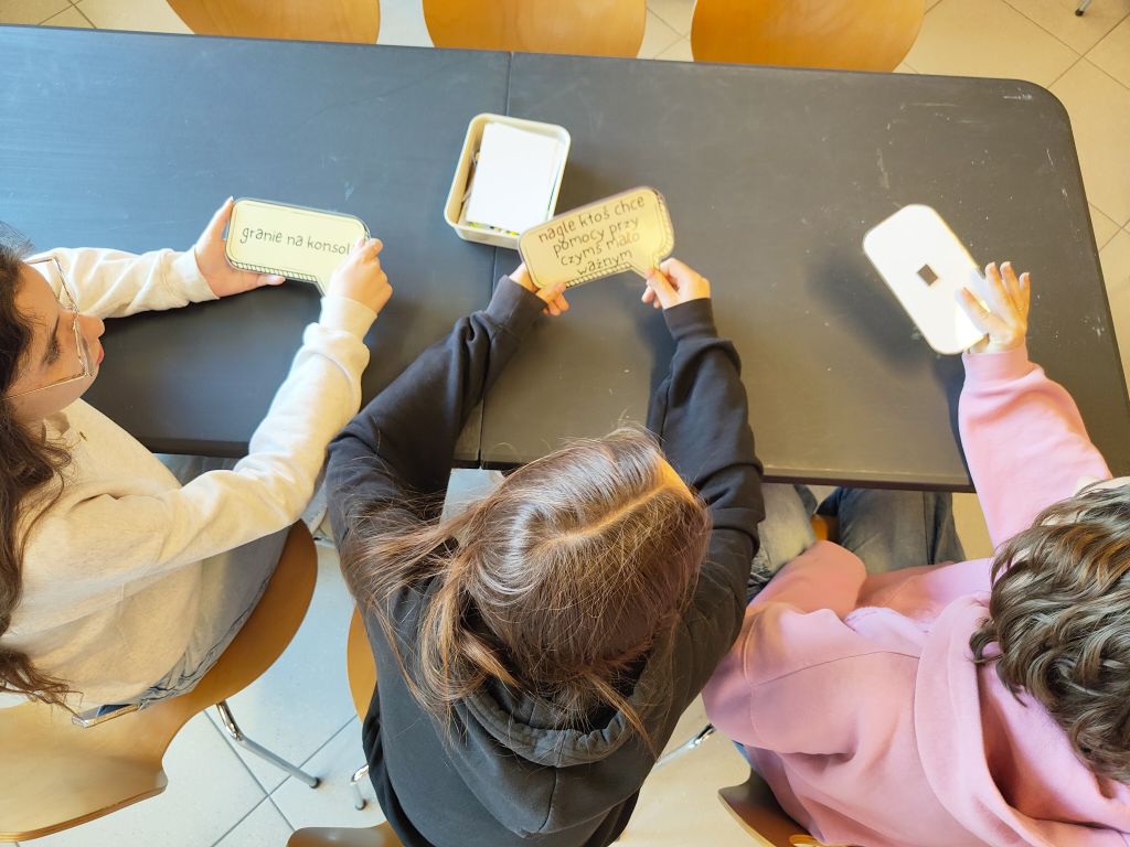 Trzy osoby siedzące przy stole trzymają papierowe dymki z napisami. Widok z góry pokazuje ich głowy i ręce operujące materiałami dydaktycznymi.