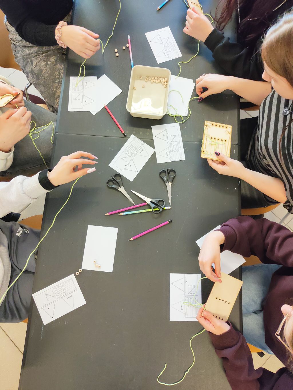Zbliżenie na dłonie uczestników warsztatów zajętych pracą manualną. Na środku stołu stoi pojemnik z drewnianymi koralikami, a obok leżą gotowe elementy do montażu.