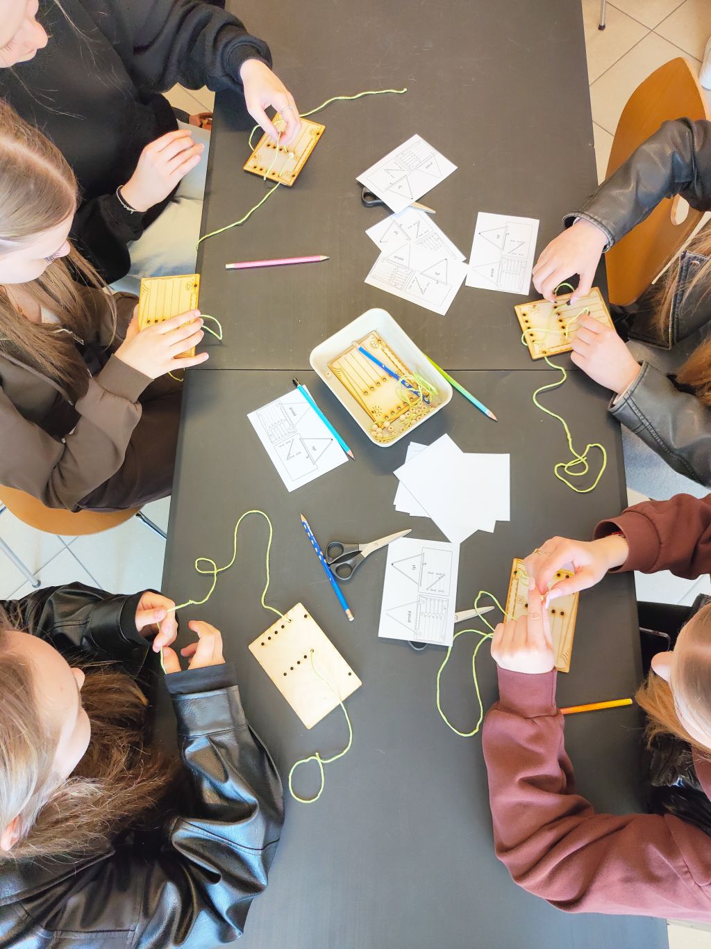 Widok z góry na młodzież pracującą przy stole. Uczestnicy przewlekają jaskrawozielone sznurki przez otwory w drewnianych tabliczkach. Na blacie leżą nożyczki, kredki i schematy pomocnicze.