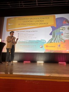 Osoba prowadząca prezentację na scenie obok slajdu o programie młodzieżowych liderów i liderek.