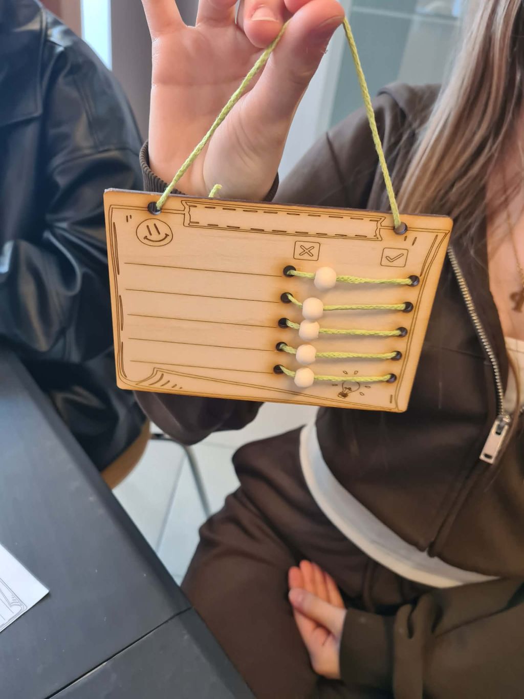 Osoba trzyma w dłoni gotową drewnianą tabliczkę z wygrawerowanym uśmiechem i żarówką, przez którą przewleczone są zielone sznurki z koralikami-suwakami.