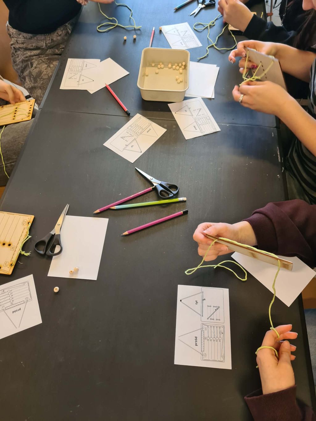 Zbliżenie na blat stołu, na którym leżą sznurki, nożyczki, koraliki i instrukcje. Dłonie uczestników pracują nad drewnianymi tabliczkami.