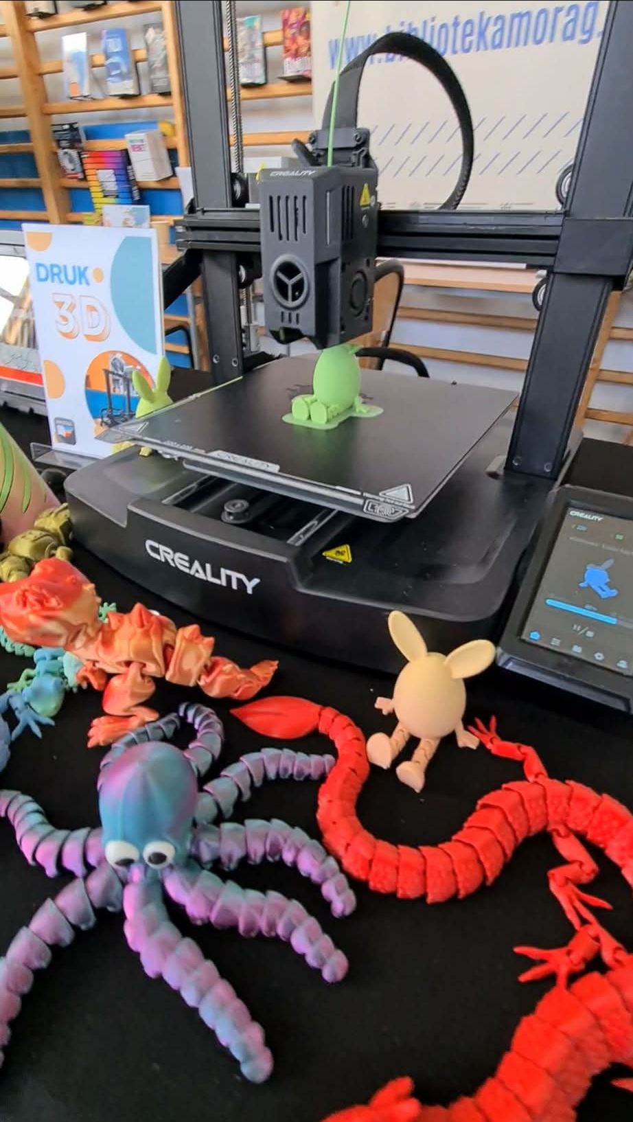 Drukarka 3D podczas pracy przy stoisku biblioteki, obok kolorowe figurki wydrukowane w technologii 3D.