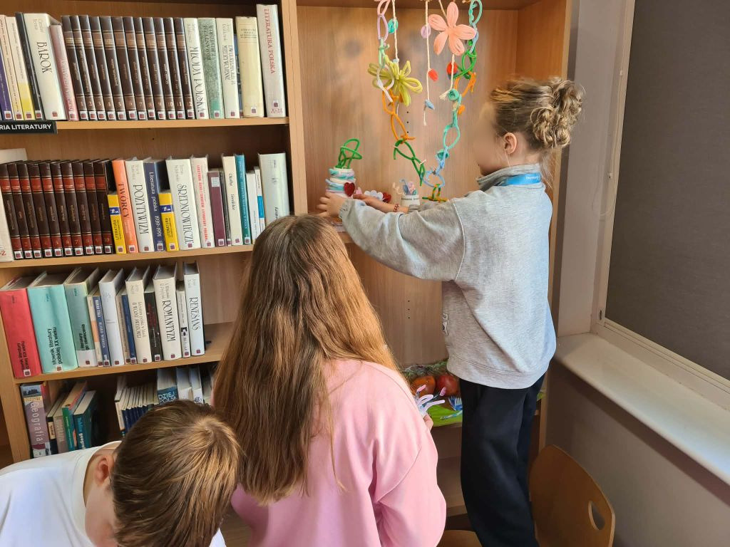 Dziecko ustawia na półce regału z książkami wykonaną własnoręcznie ozdobę z kubeczka i kolorowych drucików kreatywnych.