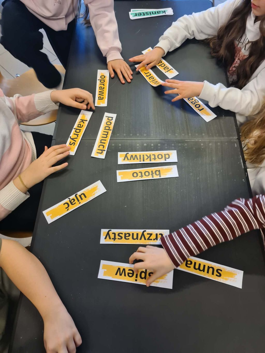 Dłonie kilku osób przesuwające po czarnym blacie żółte etykiety z polskimi wyrazami w ramach ćwiczenia grupowego.