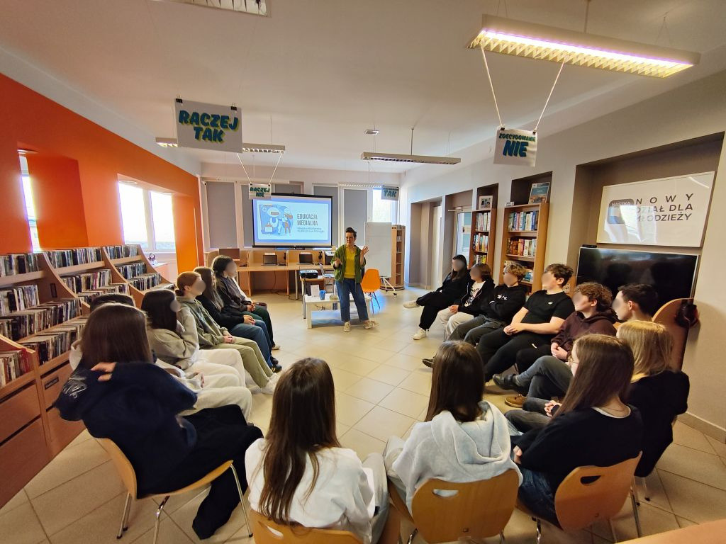 Prowadząca zajęcia kobieta stoi przed ekranem z napisem „EDUKACJA MEDIALNA” i przemawia do młodzieży siedzącej w półkolu. Na ścianie po prawej znajduje się tablica z napisem „NOWY DZIAŁ DLA MŁODZIEŻY”.