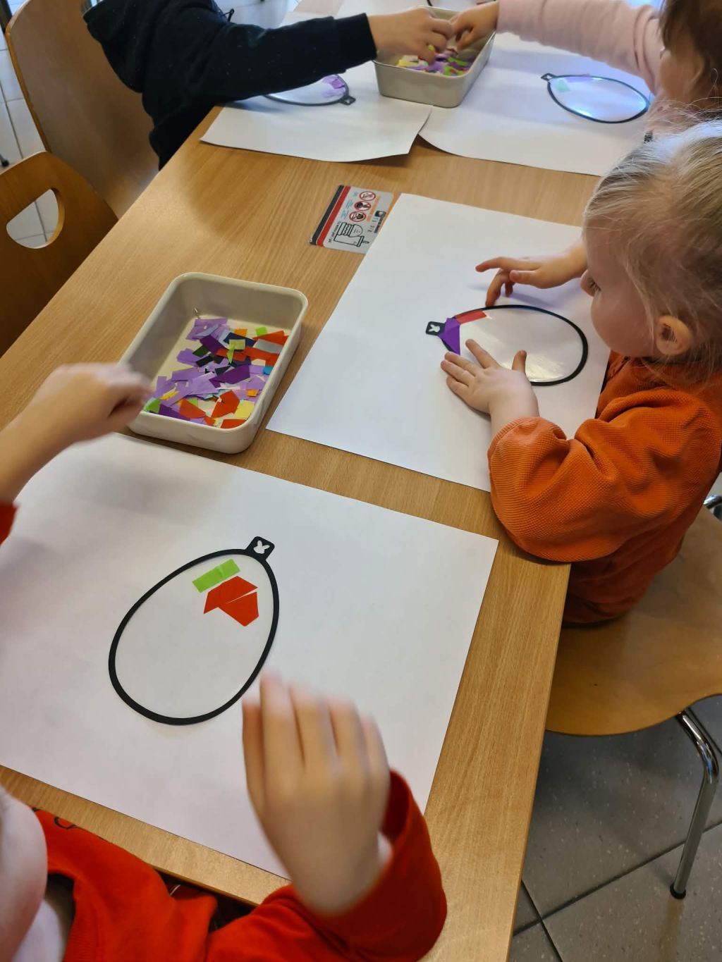 Zbliżenie na dłonie dwójki dzieci naklejających fioletowe, zielone i pomarańczowe kawałki bibuły na samoprzylepną powierzchnię wewnątrz czarnego konturu jajka.