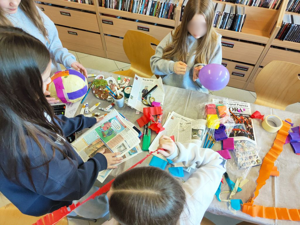 Trzy dziewczynki przy stole pracują nad przestrzennymi formami; jedna trzyma fioletowy balon, inne wycinają paski kolorowej bibuły.