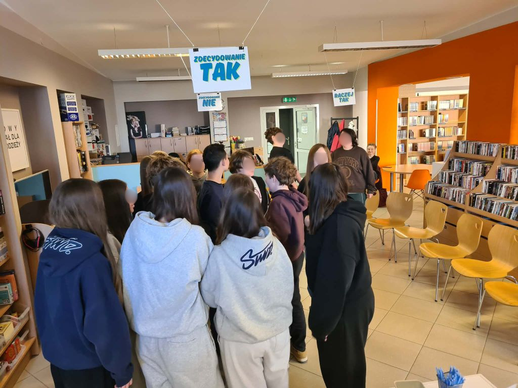 Grupa młodych ludzi stoi na środku sali bibliotecznej, pod wiszącymi u sufitu dużymi napisami „ZDECYDOWANIE TAK” oraz „ZDECYDOWANIE NIE”. Pomieszczenie jest jasne, z widocznymi w tle stanowiskami komputerowymi.