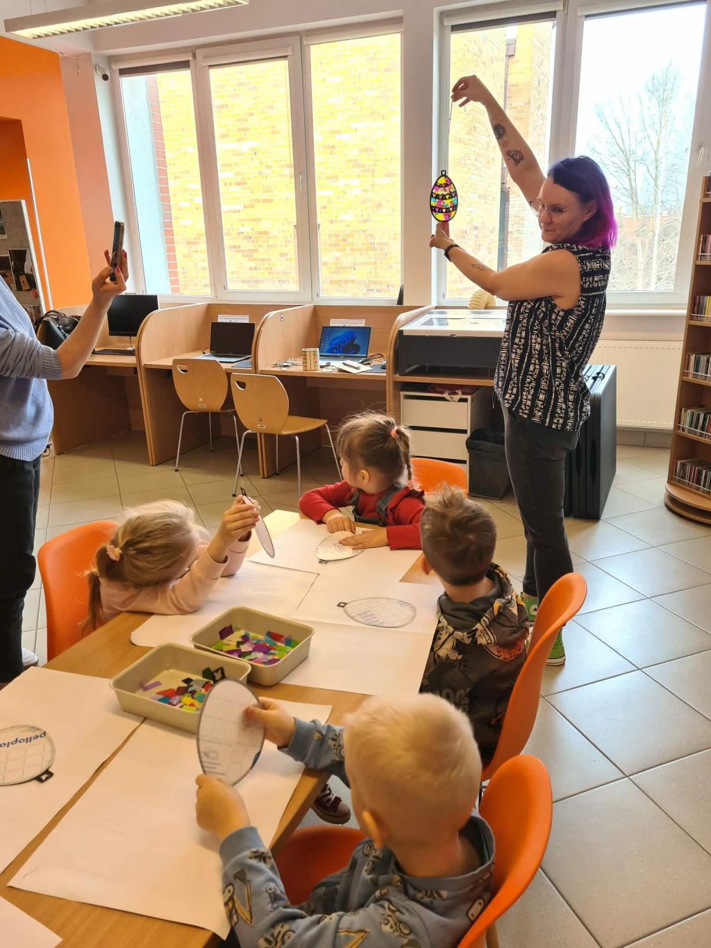 Kobieta z fioletowymi włosami trzyma w górze wyklejoną, kolorową ozdobę w kształcie jajka, pokazując ją dzieciom siedzącym przy stole. W tle widoczne są stanowiska komputerowe oddzielone niskimi ściankami.