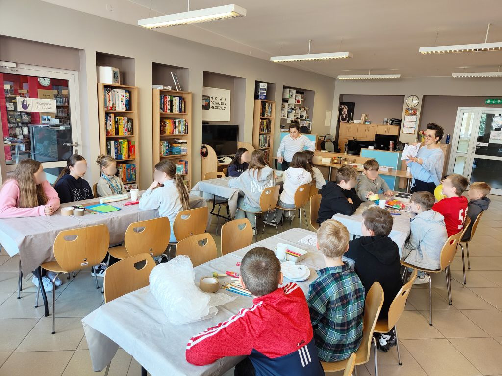 Grupa dzieci siedzi przy stołach w nowoczesnej bibliotece, słuchając instrukcji prowadzącej, która trzyma kartkę z zadaniem.