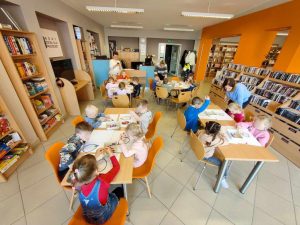 Szerokie ujęcie jasnej sali bibliotecznej z pomarańczową ścianą i regałami pełnymi książek oraz gier. Grupa kilkanaściorga dzieci siedzi przy czterech prostokątnych stołach, wyklejając kolorową bibułą kontury jajek na białych kartkach. W głębi sali stoją trzy dorosłe kobiety obserwujące pracę dzieci.