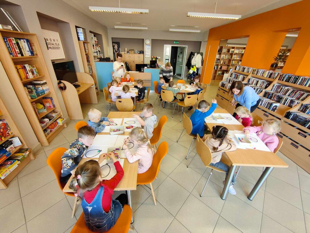 Szerokie ujęcie jasnej sali bibliotecznej z pomarańczową ścianą i regałami pełnymi książek oraz gier. Grupa kilkanaściorga dzieci siedzi przy czterech prostokątnych stołach, wyklejając kolorową bibułą kontury jajek na białych kartkach. W głębi sali stoją trzy dorosłe kobiety obserwujące pracę dzieci.