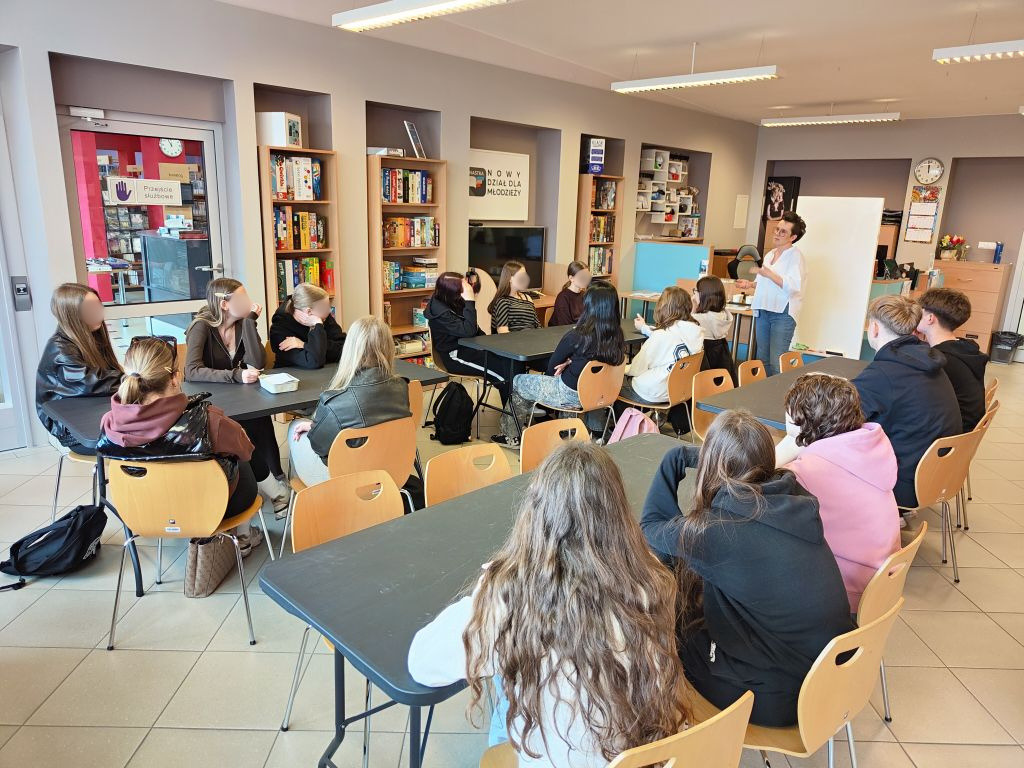 Grupa młodzieży siedzi przy czarnych stolikach w nowoczesnej bibliotece, słuchając kobiety stojącej przy białej tablicy. W tle regały z grami planszowymi