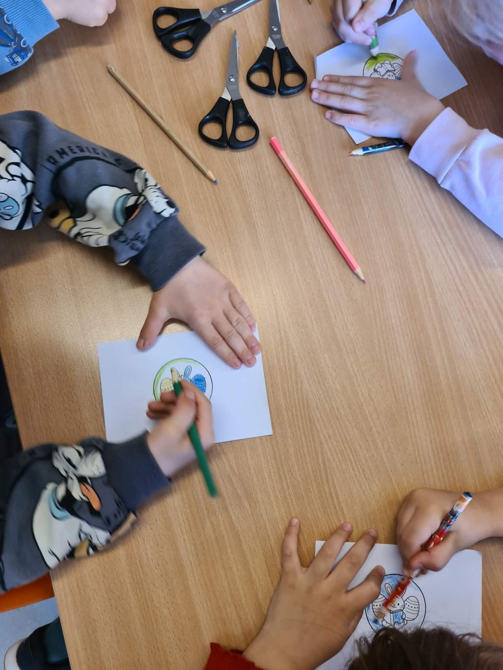 Zbliżenie na ręce trójki dzieci kolorujących kredkami małe rysunki z motywami wielkanocnymi. Na drewnianym blacie leżą również trzy pary nożyczek z czarnymi uchwytami oraz kolorowe kredki.