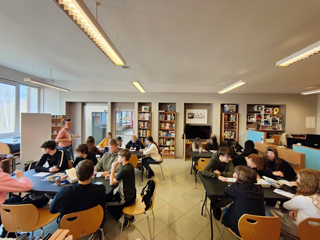 Młodzież pracuje w grupach przy stolikach w przestrzeni działu dla młodzieży w bibliotece.