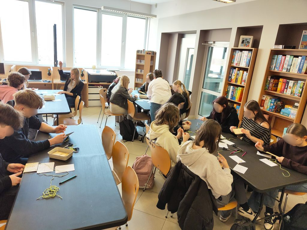 Młodzież pracuje w skupieniu przy stolikach, wykonując zadania manualne z użyciem sznurków, nożyczek i drewnianych elementów.