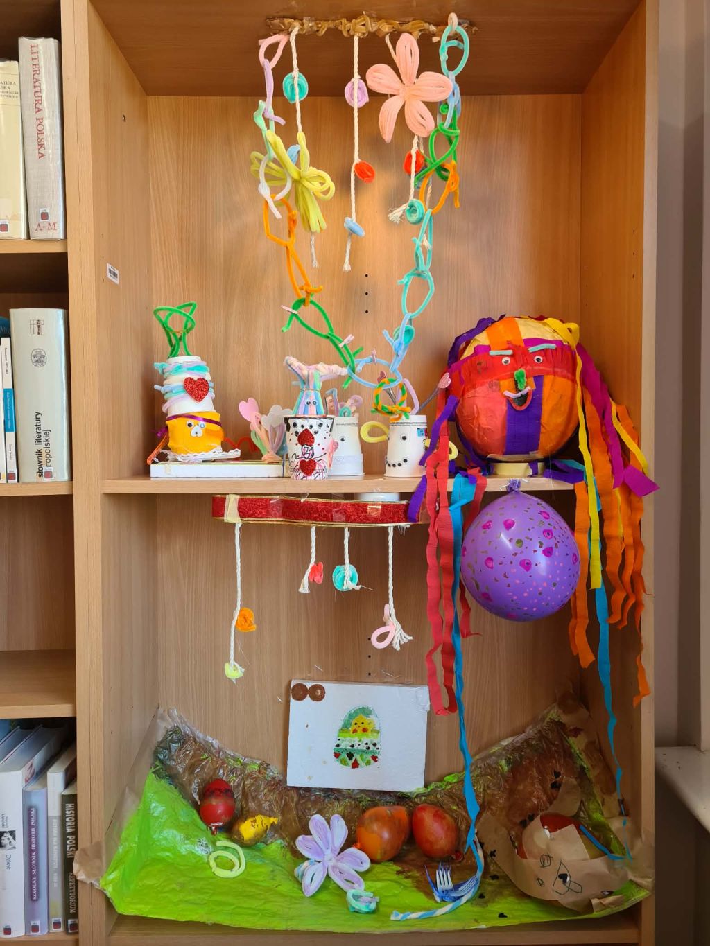Regał z książkami, na którym wyeksponowano gotowe prace: kolorowe stworki z kubeczków, postać z balonu z włosami z bibuły oraz wiszące ozdoby.