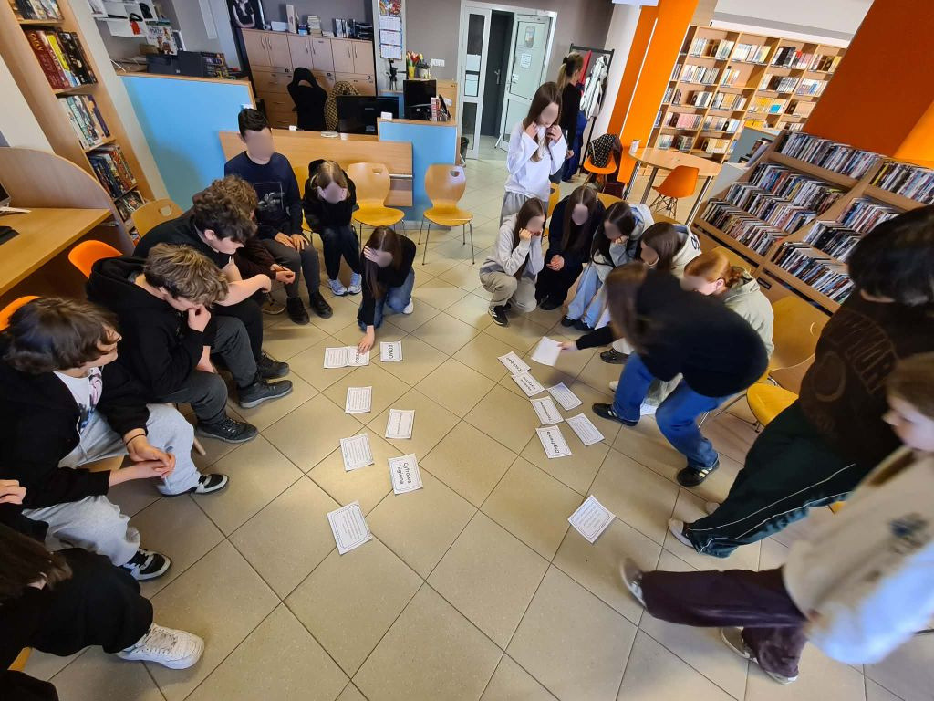 Uczniowie kucają i siedzą na podłodze wokół rozłożonych białych kartek z napisami, takimi jak „Cyfrowa higiena”, „FOMO” czy „Wstęp”. Wspólnie analizują treści materiałów edukacyjnych.