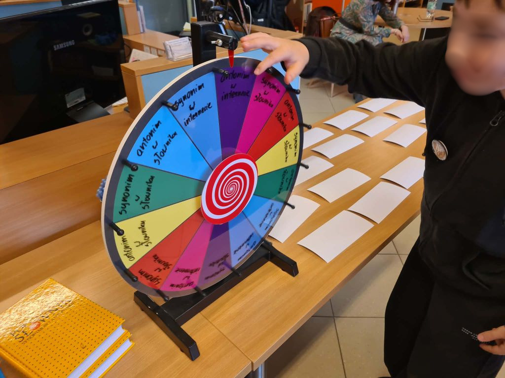 Zbliżenie dłoni dziecka obracającego kolorowe koło fortuny z napisami stojące na drewnianym stole w bibliotece, w tle leżą białe karty.