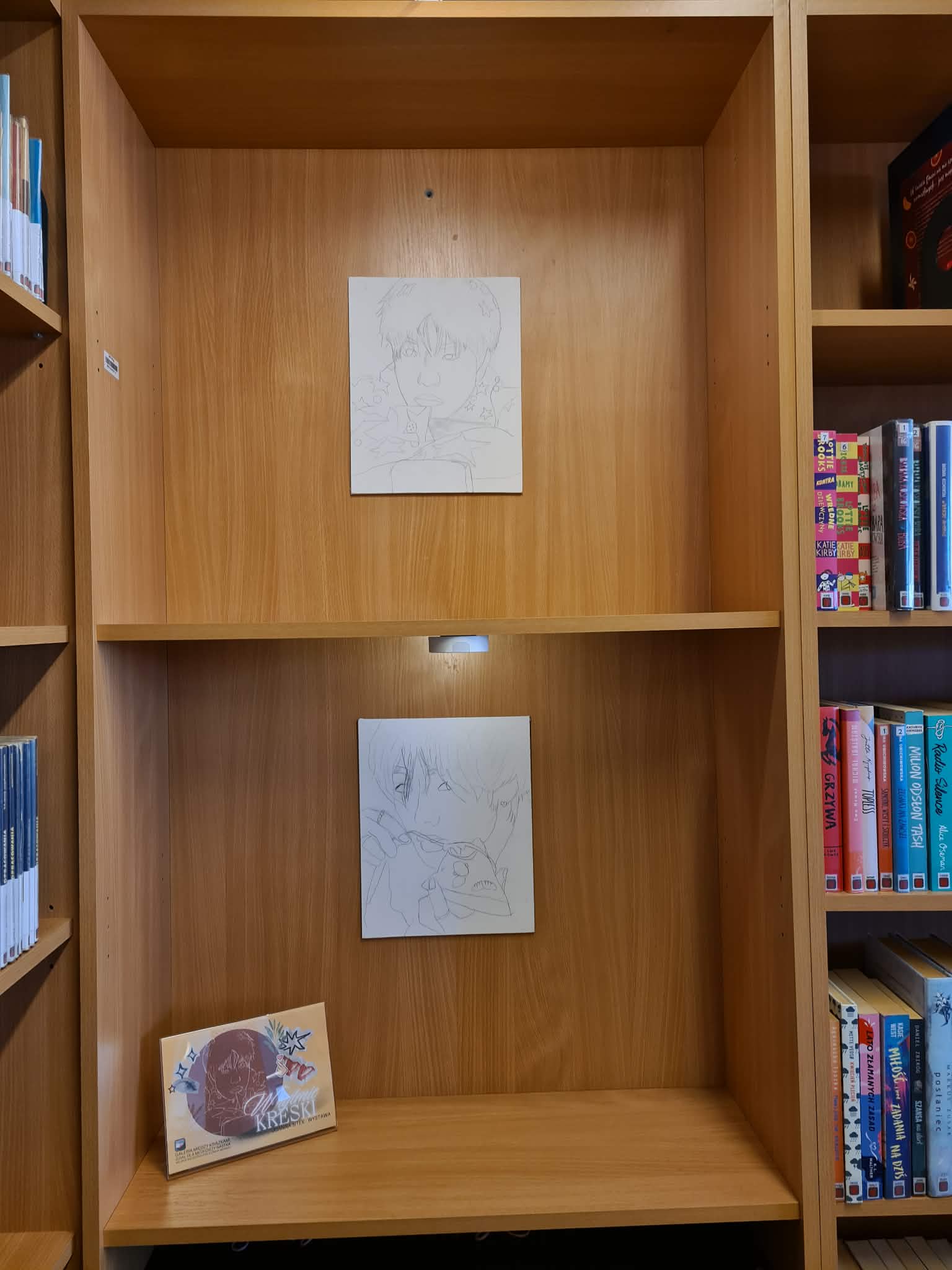 Dwie prace rysunkowe wyeksponowane na drewnianym regale bibliotecznym, obok książek i tabliczki wystawy „W rytmie kreski