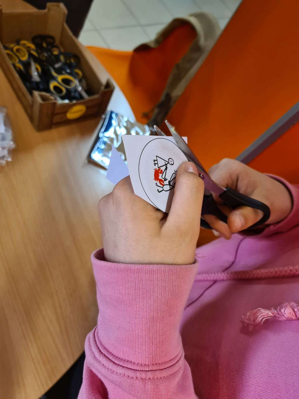 Dłonie dziecka wycinają nożyczkami okrągłą grafikę z papieru, przygotowywaną do wykonania przypinki.