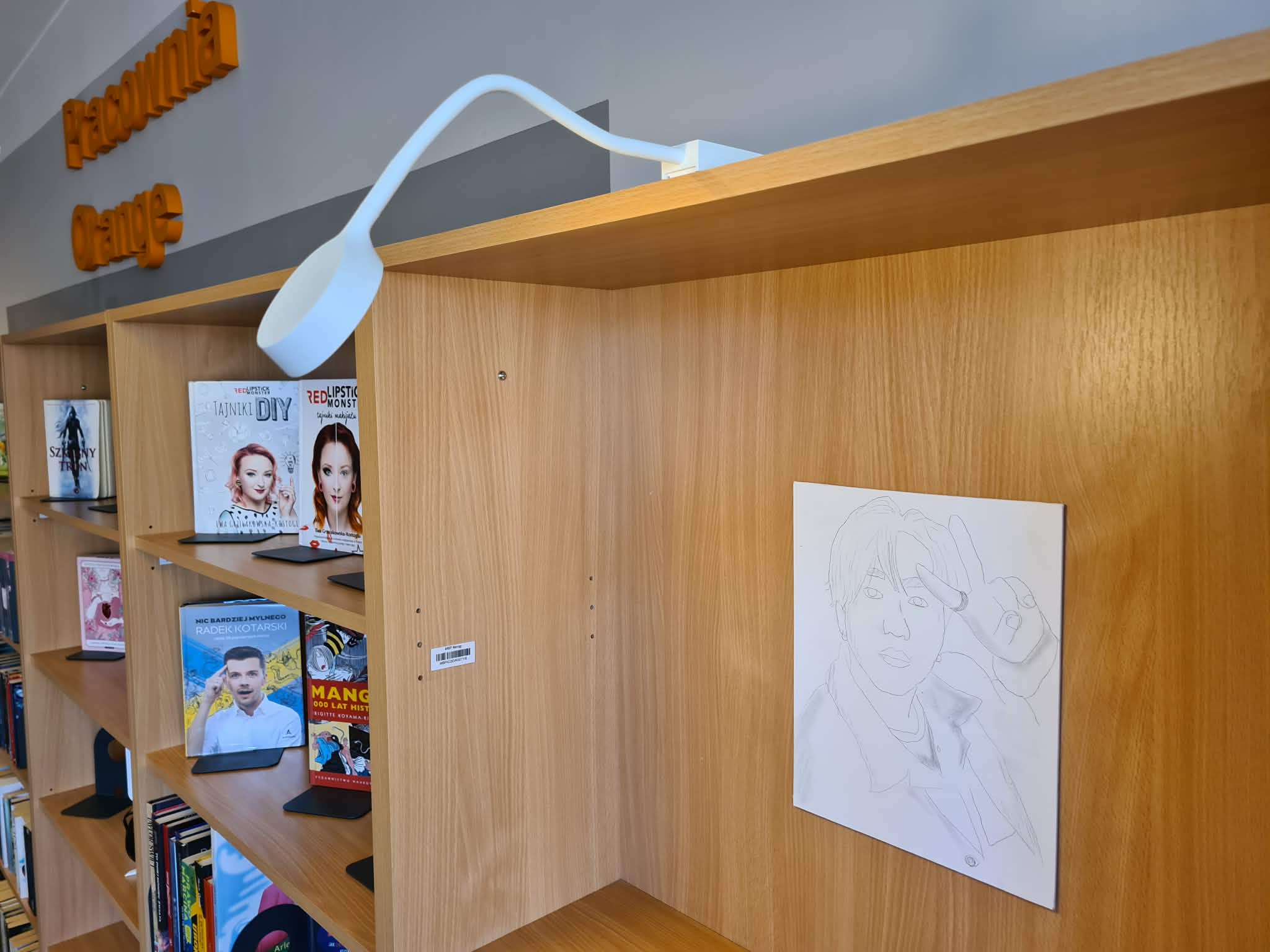 Regał biblioteczny w Pracowni Orange z rysunkiem portretowym zawieszonym na drewnianej ściance oraz lampką zamocowaną nad półką