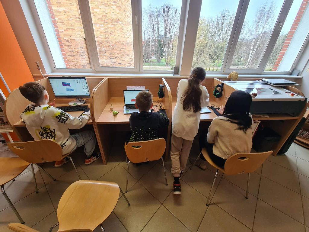Dzieci pracują przy komputerach ustawionych przy oknie w bibliotece.