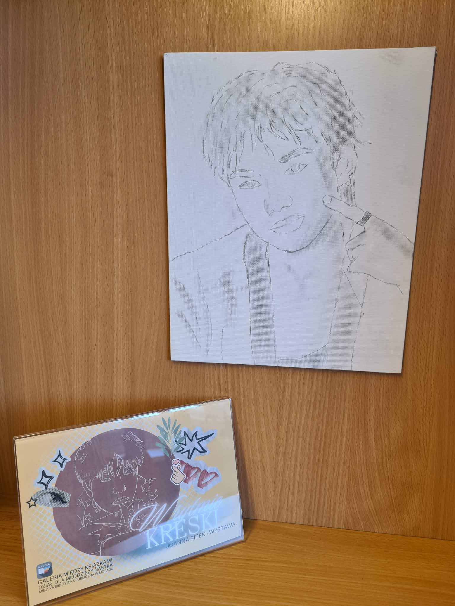 Portretowy szkic na płótnie zawieszony na drewnianej ściance regału, poniżej tabliczka wystawy „W rytmie kreski