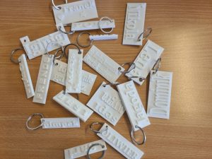 Białe breloki z imionami i napisami wykonane techniką druku 3D, ułożone na drewnianym blacie.