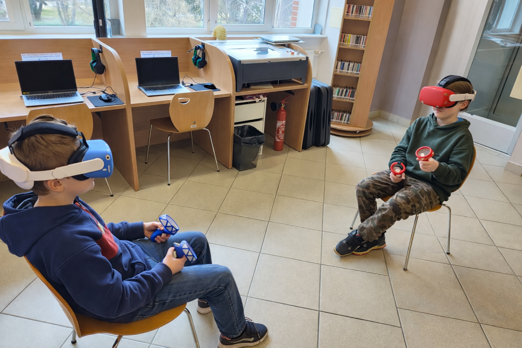 Dwoje dzieci w goglach VR siedzi na krzesłach naprzeciwko siebie w sali edukacyjnej. Jedno ma niebieską osłonę na goglach, drugie czerwoną. W tle widać biurka z laptopami i regał z płytami.
