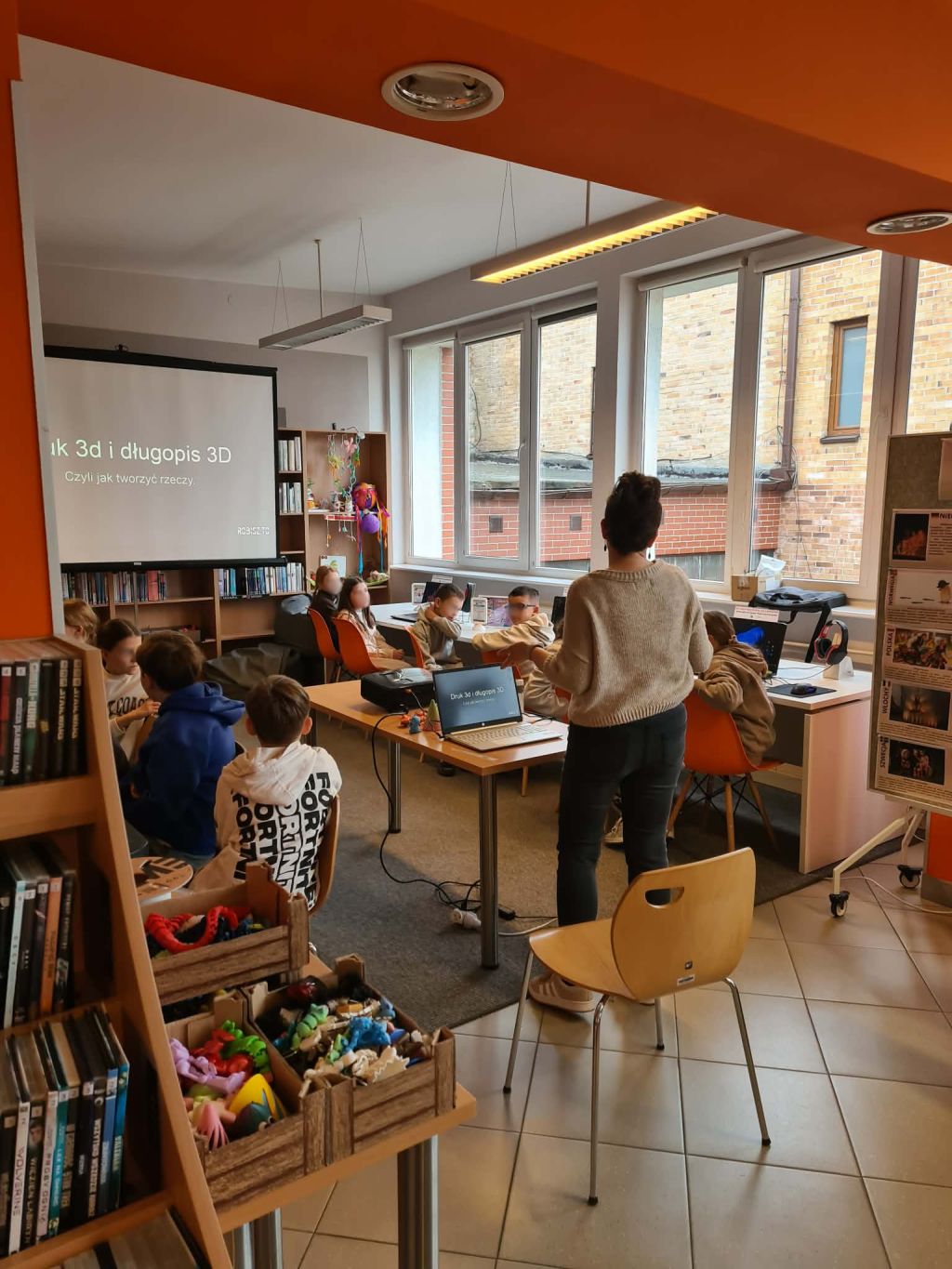 Warsztaty o druku 3D w bibliotece; prowadząca omawia temat, a dzieci siedzą przy stołach i komputerach.