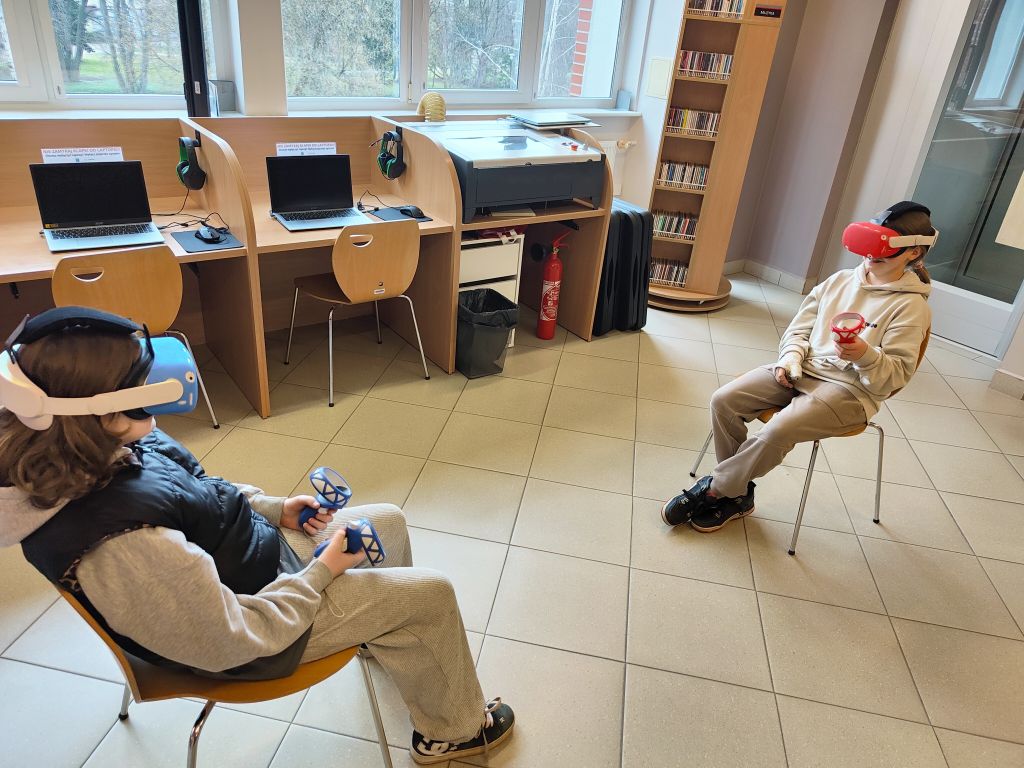 Dwoje dzieci w goglach VR siedzi na krzesłach naprzeciwko siebie w sali edukacyjnej. Jedno ma niebieską osłonę na goglach, drugie czerwoną. W tle widać biurka z laptopami i regał z płytami.